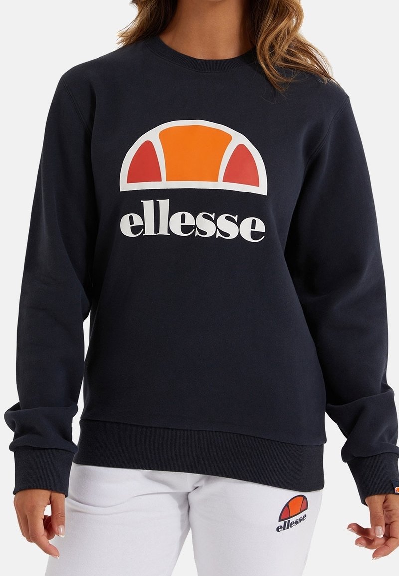 Personne portant un sweat-shirt noir ellesse avec un logo orange et rouge et un pantalon blanc ellesse avec un logo assorti sur la partie supérieure de la cuisse gauche.