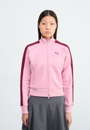 A young woman stands against a plain background, wearing a pink Puma zip-up jacket with maroon stripes and a grey pleated skirt.