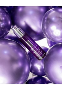 Botella de suero púrpura con un dosificador metálico, colocada entre globos púrpuras brillantes. La botella tiene un diseño elegante, acentos plateados y etiquetado claro.
