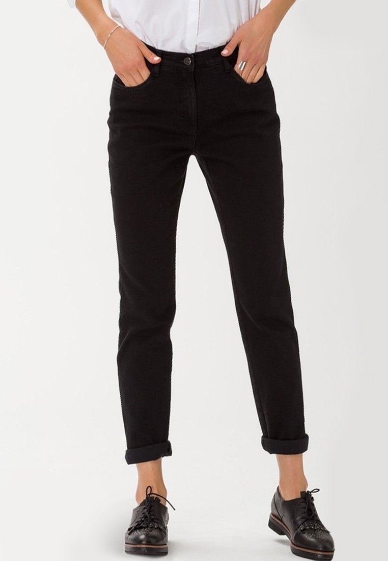 Pantalones de mezclilla negros de corte ajustado, con puños enrollados, bolsillos frontales y cierre de botón. Llevados con zapatos negros de cordones y una camisa blanca.