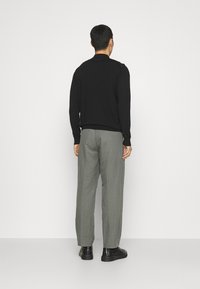 Svart stickad tröja med lång ärm, kombinerad med ljusgrå byxor och svarta skor. Utsikt bakifrån mot en enkel vit bakgrund.