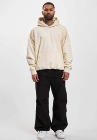 Crème kleurige hoodie met een kangoeroe zak, relaxte pasvorm en capuchon met trekkoorden, gecombineerd met zwarte losse broek en witte sneakers.
