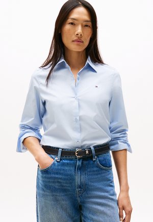 Kvinde iført en lyseblå skjorte med knapper, ærmerne rullet op, og blå jeans, med hånden i lommen, stående foran en hvid baggrund.