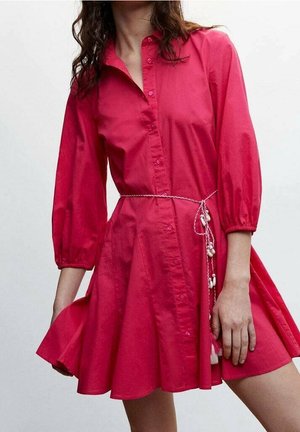 Žena v jasně růžových šatech na knoflíky se tříčtvrtečními rukávy a tenkým pleteným pásem se střapci uvázaným v pase.