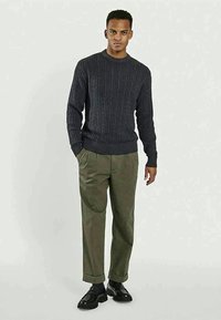 Maglione grigio scuro a trecce con scollatura rotonda abbinato a pantaloni verde oliva. Scarpe nere completano l'outfit, con i polsini risvoltati.