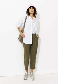 Chemise blanche à boutons, pantalon vert olive, baskets argentées et sac à imprimé léopard. Cheveux bouclés et casquette rose pâle complètent le look.