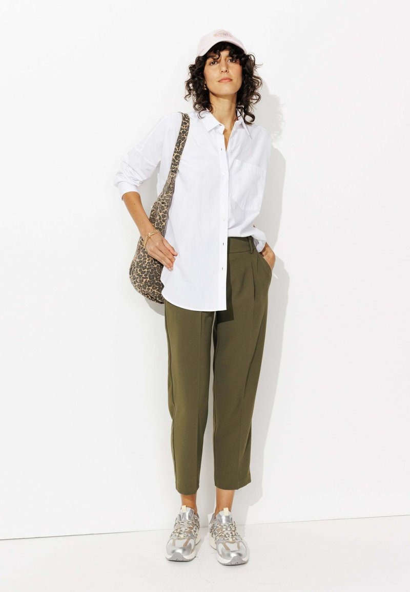 Chemise blanche à boutons, pantalon vert olive, baskets argentées et sac à imprimé léopard. Cheveux bouclés et casquette rose pâle complètent le look.