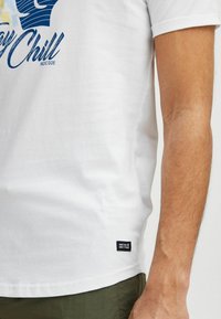 Camiseta blanca de algodón con estampado gráfico azul, mangas cortas y una etiqueta de tela negra en el dobladillo. Combinada con pantalones cortos de tela verde.
