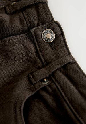 Tumepruuni teksariie, millel on metallist nööp, millele on graveeritud "NEXT DENIM". Sisaldab külgsuunda ja riidist silmust.