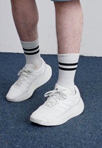 Hvite joggesko med strukturert design og snøring, parret med hvite sokker med to svarte striper. Bakgrunnen er blått teppe.