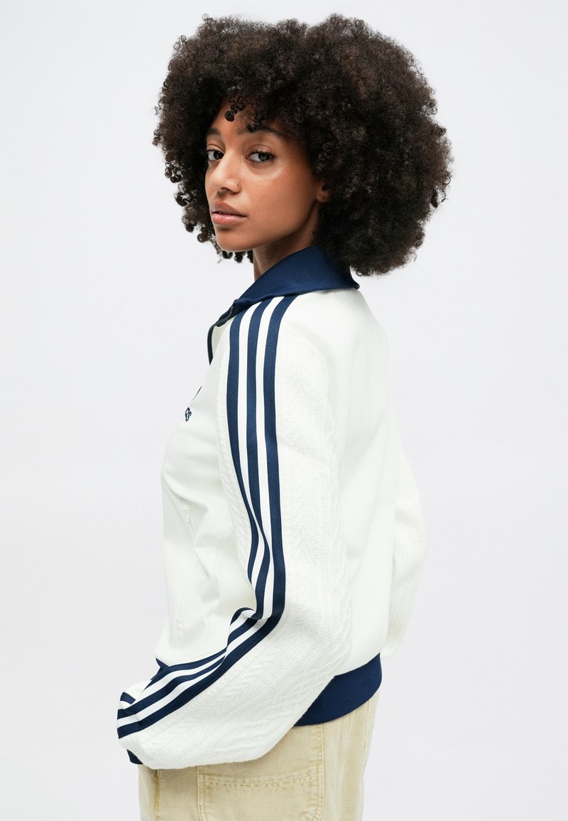 White jacket with a navy collar and three navy stripes on each sleeve. Features textured fabric on the sleeves and a zip closure.