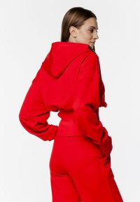 Czerwony krótki hoodie o teksturowanym wykończeniu, z dużym kapturem i luźnymi rękawami. Dopasowany z czerwonymi spodniami, tworzy spójną stylizację.