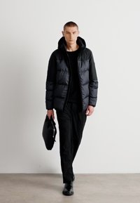 Čierna puffa bunda s kapucňou, hladká textúra, predný zips, nosená cez čierny top a nohavice, doplnená čiernou taškou a topánkami.