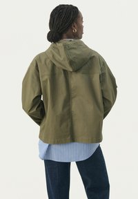 Jachetă cu glugă de culoare verde măsliniu, cu o croială lejeră, având un design drept și cusături vizibile. Sub aceasta, este stratificat un tricou în dungi albastre și albe.