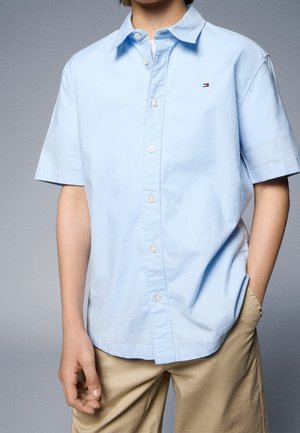 Lyseblå kortærmet skjorte i bomuld med knapper, som har en brystlomme og et lille logo på brystet, kombineret med beige bukser.