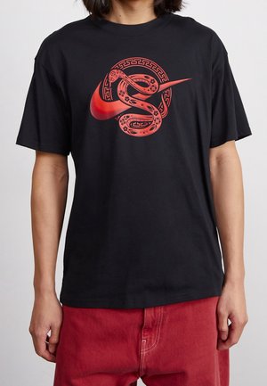 Personne portant un t-shirt noir avec un serpent rouge enroulé autour du logo Nike Swoosh et un pantalon rouge sur un fond uni.
