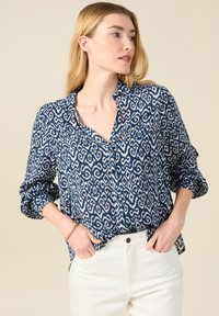 Camicia blu navy con bottoni e motivo bianco a spirale, maniche lunghe arrotolate ai polsini e vestibilità comoda. Indossata con jeans bianchi.