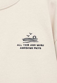 Un tissu crème présente une scène de plage brodée en bleu marine, avec des vagues, un soleil et deux oiseaux, accompagnée du texte "TOUT ÇA ET ENCORE PLUS DE JOURS GÉNIAUX."