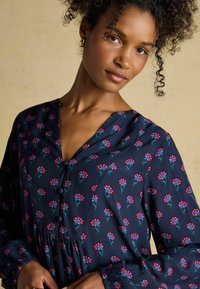 Jūras zila blūze ar ziedu rakstu, ar V veida izgriezumu, pogu aksesuāriem un elastīgām piedurknēm. Mīksta auduma blūze ar uzturētu vidukļa dizainu.