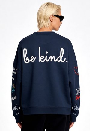 Persona con capelli biondi corti e occhiali da sole indossa una felpa blu navy con "be kind." sul retro e varie grafiche sulle maniche.