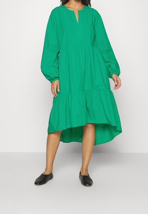 Femme portant une robe midi verte à manches longues, à ourlet haut-bas, associée à des chaussures plates noires sans lacets, debout devant un fond gris clair.