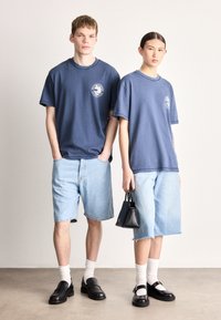 Vienodo stiliaus tamsiai mėlyni medvilniniai marškinėliai su apvaliu kaklu ir logotipu, derinami su šviesiais džinsiniais šortais. Juodi batai ir mažas juodas rankinukas su tekstūruotu paviršiumi.