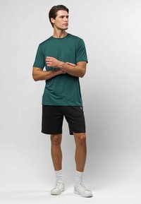 Tričko s krátkým rukávem v tmavě modrozelené barvě, s kulatým výstřihem, spárované s černými šortkami. Model na sobě má bílé tenisky a bílé žebrované ponožky.