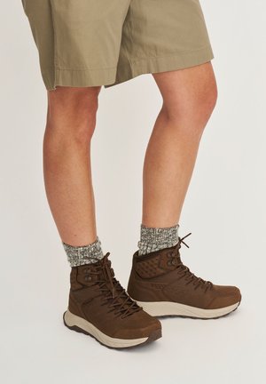 Onderbenen in khaki shorts, grijze gebreide sokken en bruine hoge wandelschoenen met witte zolen, staand op een lichte achtergrond.