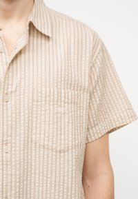 Μπεζ πουκάμισο με κοντά μανίκια, κουμπιά εμπρός, κάθετες ρίγες και τσέπη στο στήθος, φορεμένο από ένα άτομο.