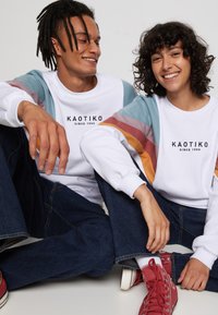 Hvid sweatshirt med multifarvede ærmer (lyseblå, rust og sennep), med mærket "KAOTIKO" trykt i sort. Denim jeans og røde sko.