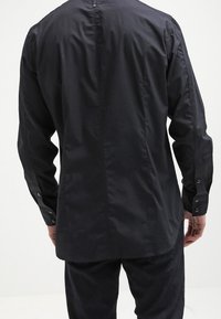 Maglietta nera a maniche lunghe con una texture liscia, polsini con bottone e una cucitura sulla schiena. Progettata con una vestibilità su misura e dettagli minimi.