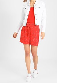 Vit denimjacka, röd tanktop, röda shorts med blommönster, knytning i midjan och vita sneakers med röda accents. Slät textur, avslappnad design.