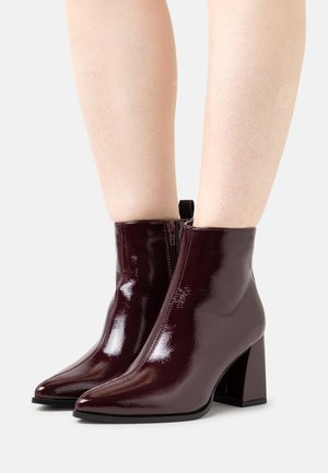 Błyszczące ciemne bordowe botki na kostkę z noskami w szpic i kwadratowymi obcasami, noszone na gołych nogach na jasnym tle.