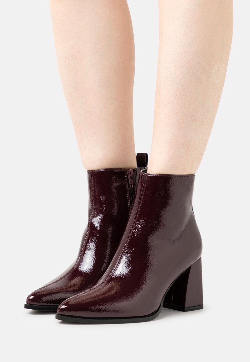 Błyszczące ciemne bordowe botki na kostkę z noskami w szpic i kwadratowymi obcasami, noszone na gołych nogach na jasnym tle.