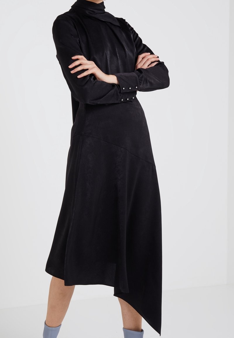 Personne portant une robe noire à manches longues avec un ourlet asymétrique et des poignets décorés de perles, debout les bras croisés sur un fond blanc.