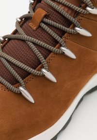 Gros plan sur une botte de randonnée en suède marron avec des lacets verts et orange à motif, passés dans des œillets métalliques sur une languette en tissu texturé.