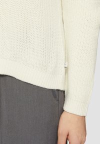 Off-white gebreide trui met een losse textuur, geribbelde zoom en boorden. Heeft een klein label aan de zijkant en wordt gedragen over grijze broeken.