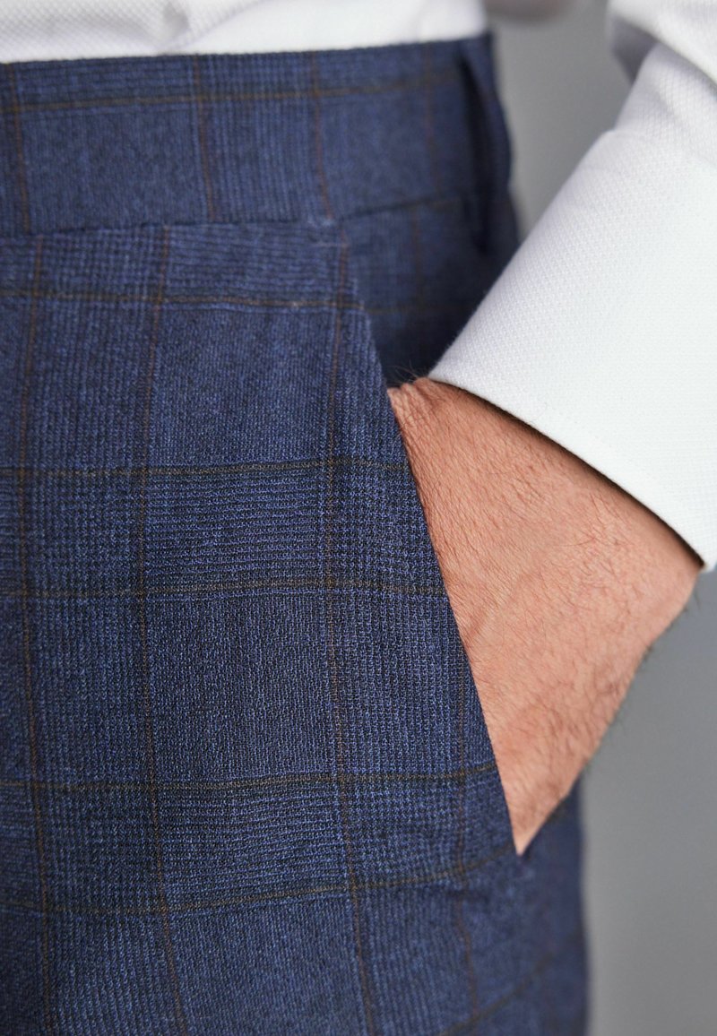Donkerblauwe geruite broek van textuurstof, met een zijzak en fijne bruine lijnen die het ontwerp accentueren. Witte mouw van een overhemd zichtbaar.