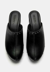 Zwarte leren klompen met een gladde afwerking, voorzien van een rij zilveren studs langs de bovenrand en een gesloten achterkant voor extra ondersteuning.