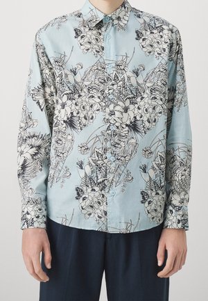 Chemise bleu clair boutonnée avec des motifs floraux détaillés en noir et blanc portée avec un pantalon foncé.