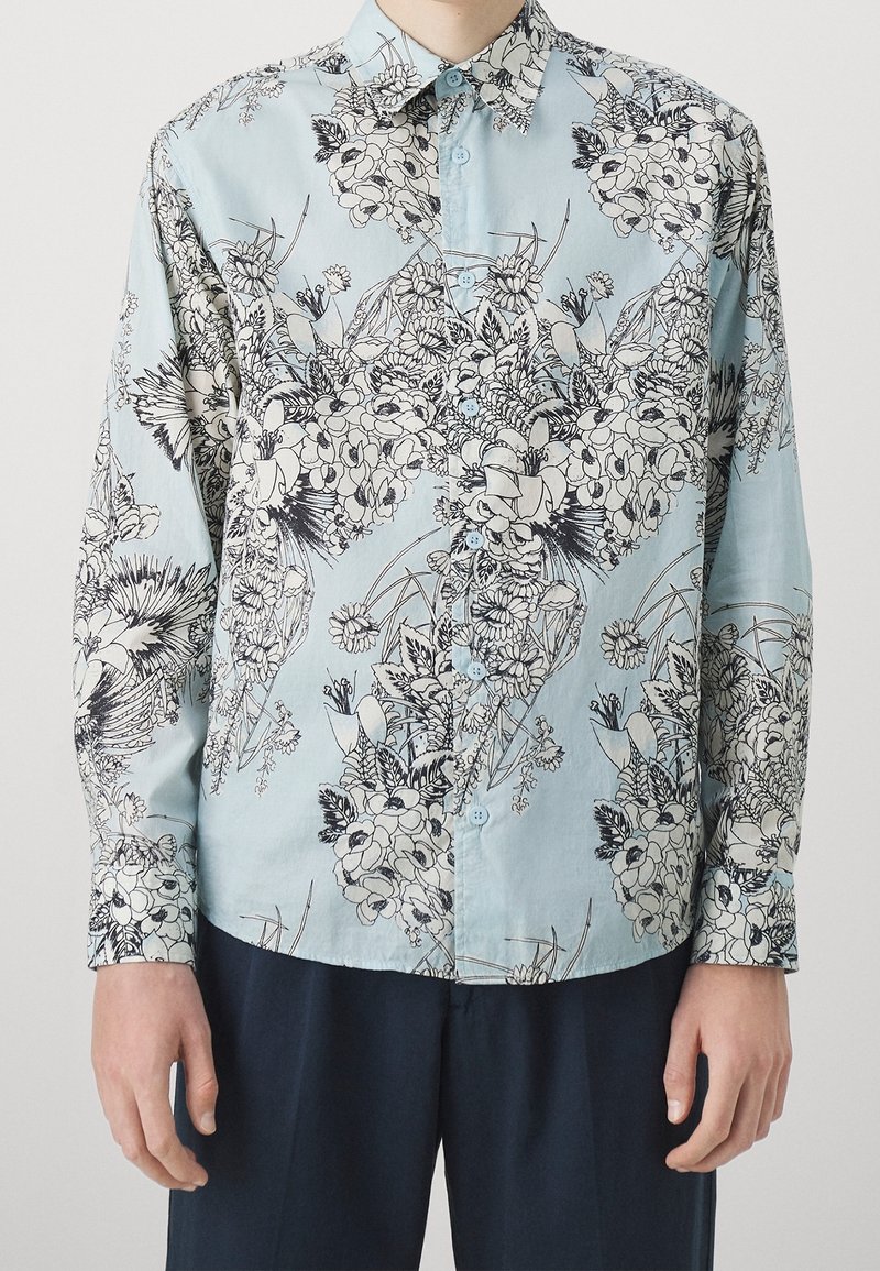 Chemise bleu clair boutonnée avec des motifs floraux détaillés en noir et blanc portée avec un pantalon foncé.