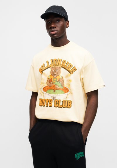 Κίτρινη βαμβακερή μπλούζα με εκτυπωμένο σχέδιο αρκούδας και κείμενο «Billionaire Boys Club». Μαύρη βερμούδα με πράσινο λογότυπο στο πλάι.