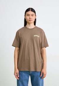 Barna pamut póló rövid ujjakkal, bal mellen világoszöld "DICKIES" logóval. Kék farmernadrággal párosítva.