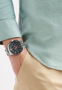 Orologio in metallo argentato con quadrante nero, accenti bianchi e blu, e bracciale in acciaio inossidabile. Indossato su un polso con una camicia azzurro chiaro.