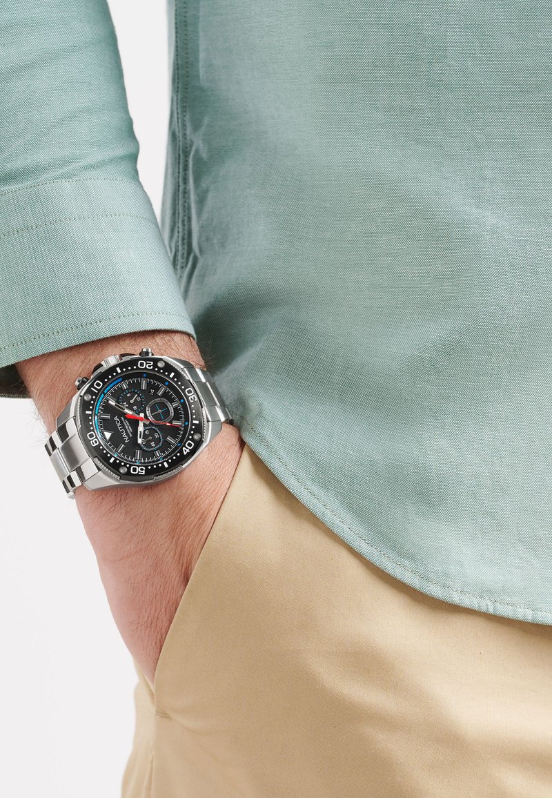 Orologio in metallo argentato con quadrante nero, accenti bianchi e blu, e bracciale in acciaio inossidabile. Indossato su un polso con una camicia azzurro chiaro.