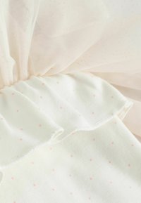 Tissu en coton crème avec de petits pois roses, présentant un bord froncé. Superposition de tulle doux et transparent dans une teinte rose clair.