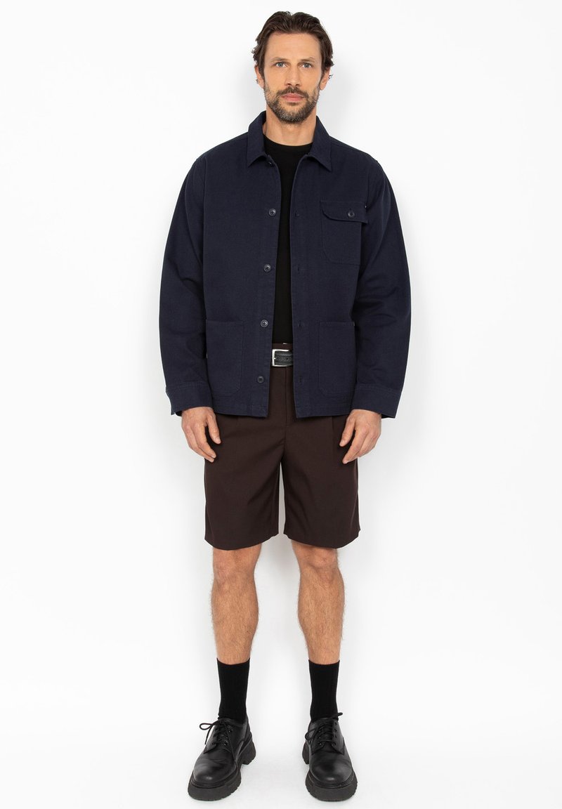 Homme debout portant une veste bleu marine, une chemise noire, un short marron, des chaussettes noires et des chaussures à lacets noires, sur fond blanc.