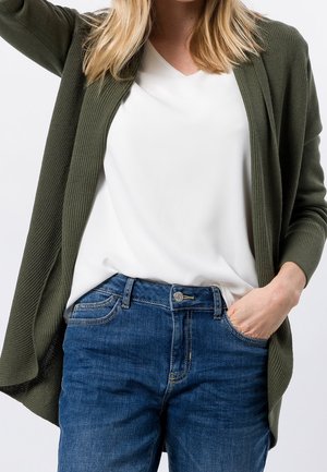 Femme portant un cardigan vert olive sur une blouse blanche avec un jean bleu, une main dans la poche, sur un fond clair uni.
