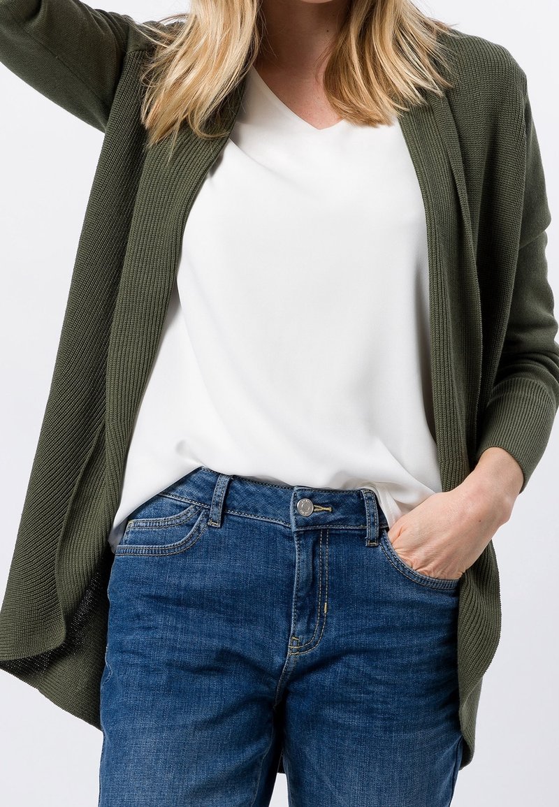 Femme portant un cardigan vert olive sur une blouse blanche avec un jean bleu, une main dans la poche, sur un fond clair uni.