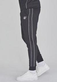 Schwarze Jogginghosen mit schmalem Schnitt, weißen Seitenstreifen und subtiler Markenkennzeichnung auf dem linken Oberschenkel, kombiniert mit weißen Sneakers.